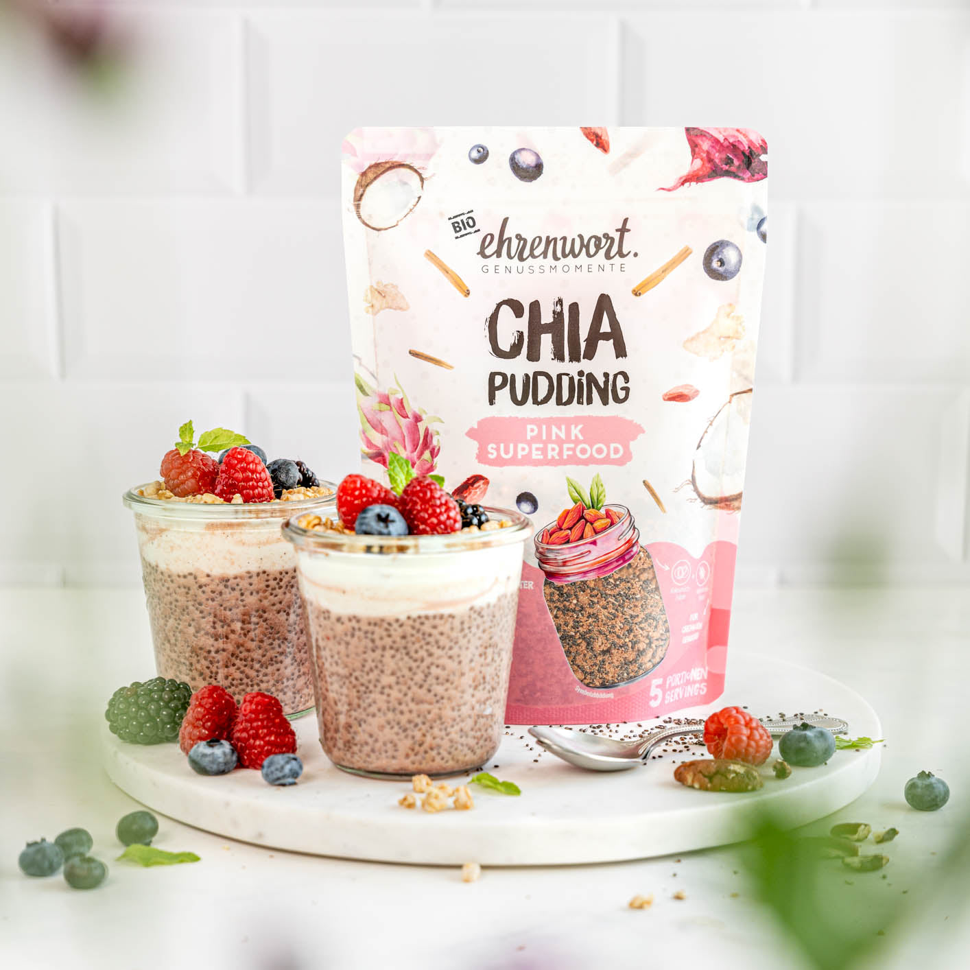 ehrenwort BIO Pink Superfood Chia Pudding Verpackung neben zubereiteter Portion in ein weißem Raum auf Präsentierteller neben Beeren und Samen